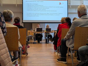 Gang zwischen Stuhlreihen, Vorne sitzen zwei Menschen an Laptops. An einer Leinwand über ihnen ist Text projiziert.