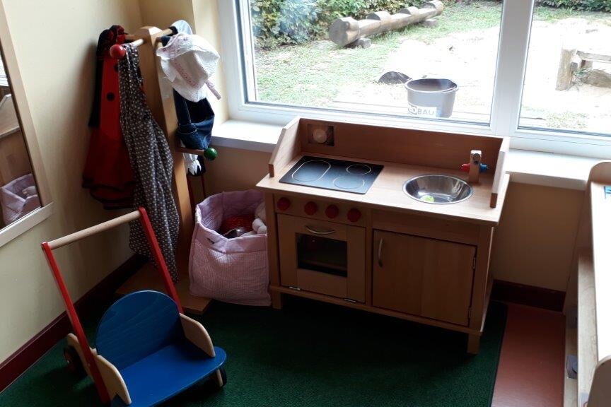 Spielküche aus Holz vor einem Fenster. In der linken Ecke ein Kindergarderobenständer.