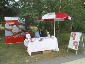 Reha Messe 2017. Eine Mitarbeiterin sitzt im Grünen an dem AWO Infostand unter einem Sonnenschirm mit AWO Logo.