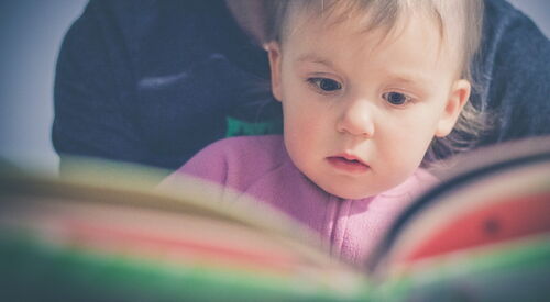 Kleinkind auf dem Schoß einer erwachsen Person, Buch ist aufgeschlagen, Kind guckt mit großen Augen.