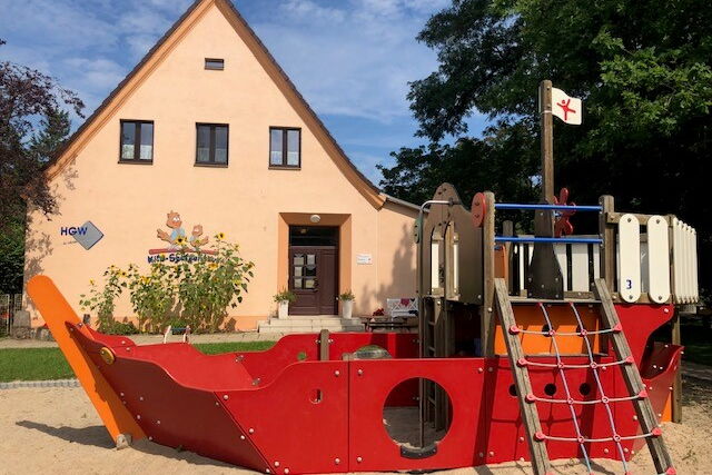 Spielplatz, Klettergerüst als Schiff mit Fahne und Steuerrad und Bullauge. Dahinter ein Haus mit Spitzdach. Das Kita-Gebäude.