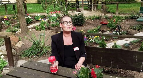 Frau sitzt an einem Tisch im Garten, Blumen blühen in den Beeten. Die Frau hat sehr kurze sunkle Haase, eine große runde, schwarz gerahmte Brille, sie trägt einen schwarzen Blazer über ein schwarzes Oberteil. Sie hält ein rote Sanduhr in der rechten Hand.