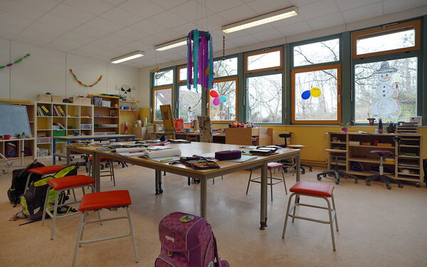 Lernzimmers. großer Tisch in der Mitte des Raumes, darauf Federtaschen, Blöcke und Hefter. Um den Tisch stehen Hocker und Schulranzen. An den Wänden stehen Regale.