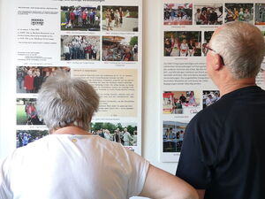 4 Menschen sehen sich große Schautafeln an einer Wand an. Chronik des Ortsbeins in Bild und Schrift.