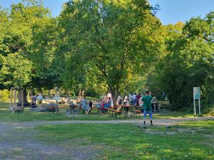 Draußen, viele schattenspende Bäume. Biertischgarnituren. Menschen in sommerlicher Kleidung. Eltern mit Kindern. Ein Spielplatz ist im Hintergrund.