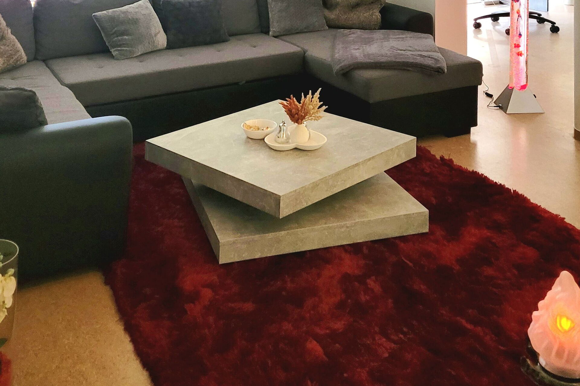 Heller großer Wohnbereich. Graues großes Ecksofa, Grünpflanze dahinter. Moderner Steintisch auf dunkelrotem Flausch-Sofa.