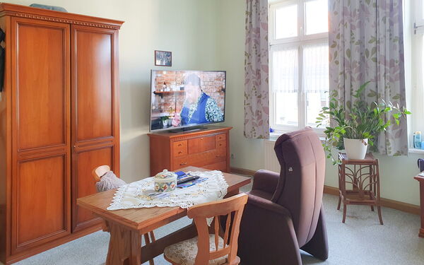 Raum mit Holzschrank und Fernsehschränkchen an der Wand. Fernseher ist an. Ein Fernsehsessel steht im Raum, daneben ein Tisch mit zwei Stühlen.