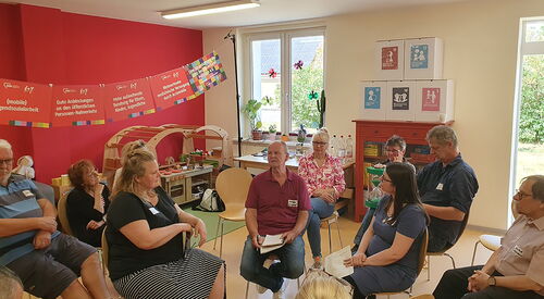 Raum mit großen Fenstern, 11 Menschen sitzen auf Stühlen, die in Kreisen angeordnet sind. In der Mitte sitzen sich zwei Frauen gegenüber ein Mann dazwischen.