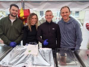 Großes Festzelt. Essen-Warmhalte Behältnisse Gastronomie. Dahinter 4 fröhliche Menschen. 3 Männer und eine Frau. Das Essen ist abgedeckt.