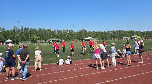 Ein Sportplatz mit Laufstrecke und Grün. Jugendliche in roten Sporttrikots auf der Grünfläche.