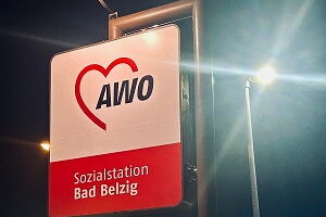 beleuchtetes Schild mit dem AWO Logo, im Hintergrund Straßenlaternen, Hauptstraße, beleuchtetes Schild von Lidl und Parkplatz von Rewe, Himmel ist dunkel