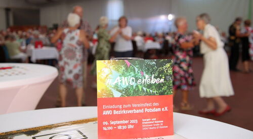 Einladung zum Vereinsfest steht an ein Glas gelehnt auf einem Tisch. Im Hintergrund fröhlich tanzende Menschen und lange Kaffeetafeln.