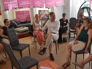 Mann sitzt im mittleren Kreis und redet lebhaft. Er trägt ein weißes langärmliges Hemd und eine helle Hose. Vor ihm 2 lehre Stühle. Im äußeren Stuhlkreis sitzen 3 Frauen und ein Mann.