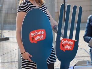 Symbolisches großes Besteck, Löffel und Gabel mit dem Logo: Schule satt!        Es wird gehalten von einer Frau mit schwarz-weiß gestreiften Kleid.