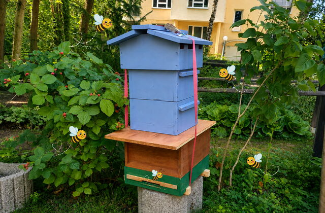 Aus Holz gebautes Haus mit mehreren Stockwerken für Bienen.   Einige virtuelle Bienen mit Gesichtern schwirren um das Häuschen.