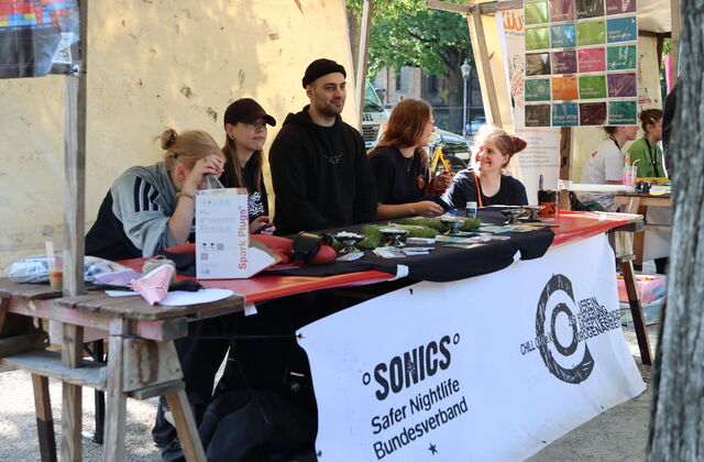 Stand mit jungen Leuten. Vorn ist ein weißes Banner befestigt. Text: Sonics Safer Nightlife Bundesverband / ChillOut.