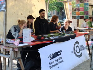 Stand mit jungen Leuten. Vorn ist ein weißes Banner befestigt. Text: Sonics Safer Nightlife Bundesverband / ChillOut.