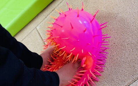 Ein Kind steht mit nackten Füßen auf einem pinken, weichen "Igelball". Am Bildrand ist eine grüne Matte zu sehen.