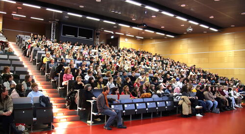 Hörsaal einer Uni. Gut besucht, fasst alle Plätze sind besetzt.