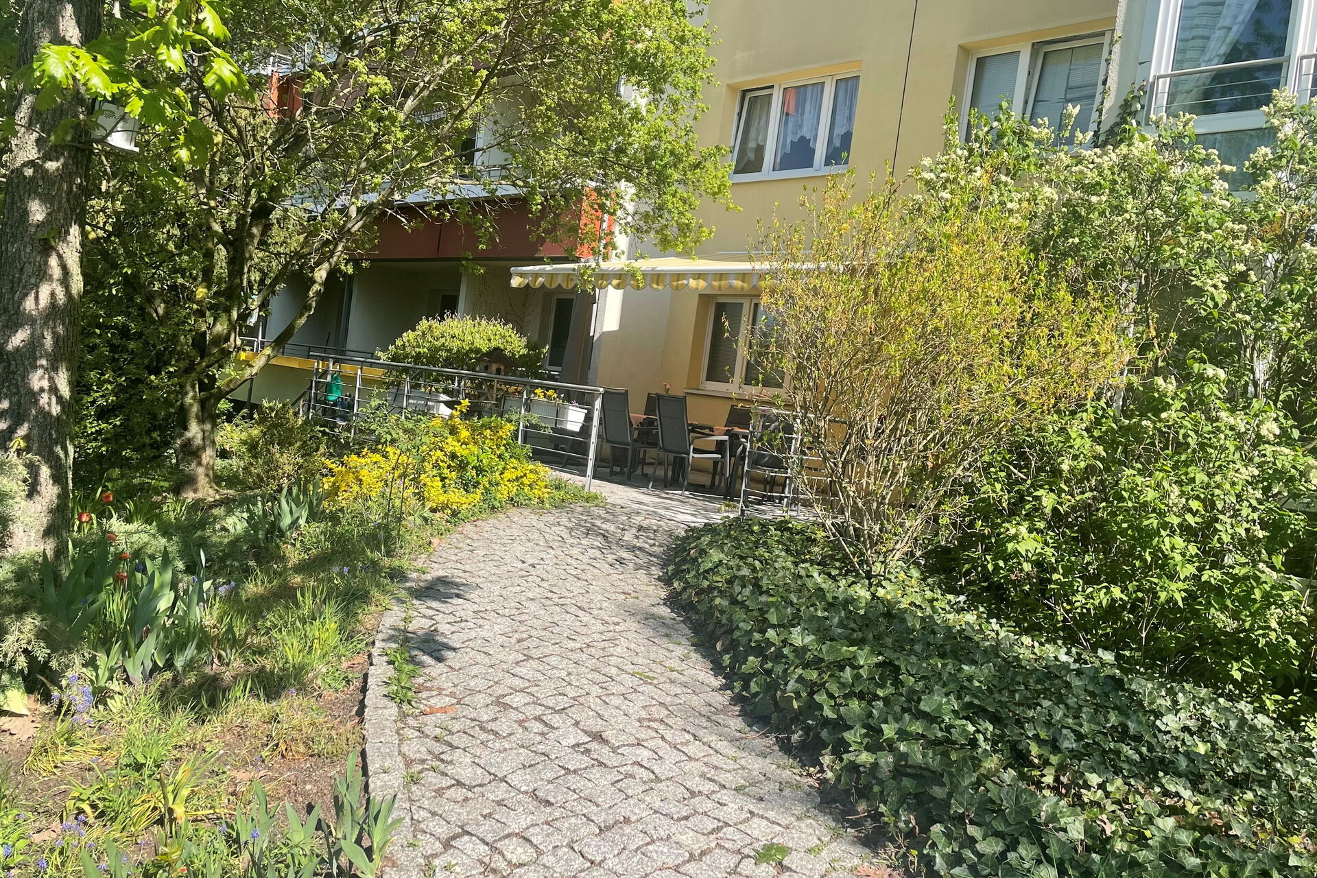 Barrierearmer Zugang über den Garten auf die Terrasse. Es stehen Stühle und Tische auf der Terrasse und eine Markise spendet Schatten.