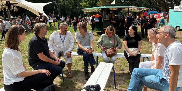 Gruppenbild, 8 Menschen sitzen auf Bierbänken in U-Form. Dahinter Festival Gelände mit Getränkewagen.