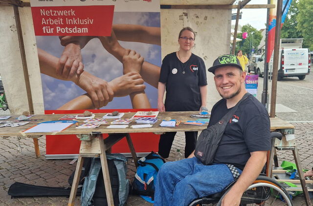 Ein Stand auf dem Fest mit einem Plakat von Netzwerk Arbeit inklusiv für Potsdam. Info-Material auf einem Tisch. Eine Frau im AWO T-Shirt steht hinter dem Tisch. Davor ein Rollstuhlfahrer im Aktivrollstuhl mit AWO T-Shirt und Base Cape.