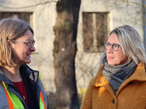 Draußen. 2 Frauen reden miteinander. Ein Mann steht rechts im Hintergrund.