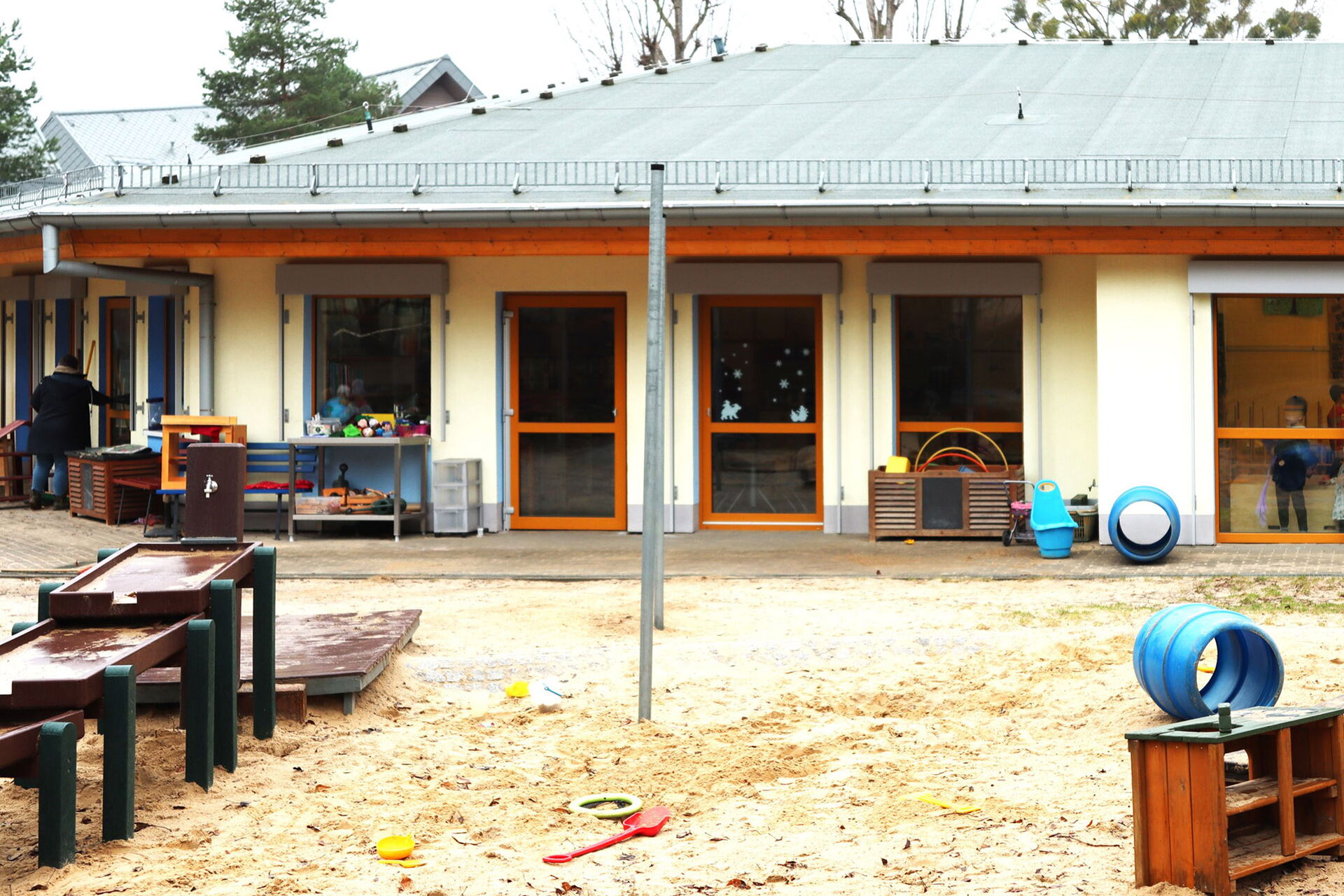 Außenansicht einer Kita. Viele Fenster. Ein Kind schaut durch ein Fenster nach draußen. Spielplatz: Sandkasten und eine Wasserpumpe.