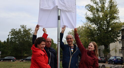 Ein Straßenschild mit einem weißen Tusch darüber. Von unten greifen 2 Frauen und 2 Männer nach dem Tusch. Die Enthüllung steht kurz bevor.