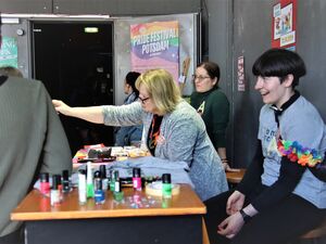 Ein Tisch von der Seite. Darauf steht Nagellack in verschiedenen Farben. Drei Frauen sind hinter dem Tisch. Es liegen auch graue T-Shirts auf dem Tisch.