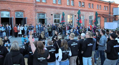 Viele Menschen vor einem rotenn Backsteingebäude. In der Mitte der Menschen sind Musikerinnen mit Gitarren. Viele Leute tragen Kleidung mit Aufdruck vom Potsdamer Kneipenchor.