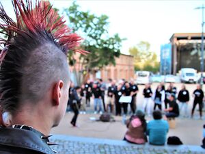 Punk mit rotem Irokesenschnitt und Nietenhalsband blickt auf einen Platz mit Menschen, größtenteils in schwarzer Kleidung die jedoch unscharf sind.