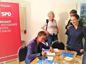 Frau sitzt an einem Tisch und signiert ihre Bücher. Frau hat kurze Haare, Brille und trägt einen lila Blazer. Drei Personen stehen am Tisch und warten. Hinter der Autorin ist ein Roll-Up der SPD Potsdam-West