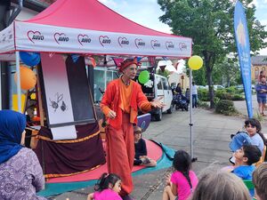 Ein farbenfroh gekleideter Mann mit Narrenmütze bespaßt Kinder und Erwachsene. Er trägt Orange-Rote Kleidung.