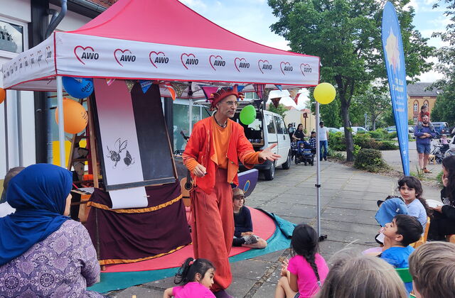 Ein farbenfroh gekleideter Mann mit Narrenmütze bespaßt Kinder und Erwachsene. Er trägt Orange-Rote Kleidung.