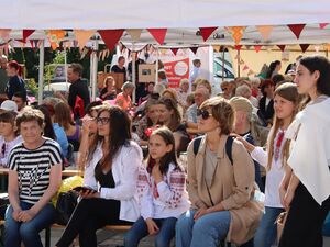 Sonnig, warm, draußen, viele Menschen sitzen auf Bänken. Auch viele Kinder. Mit bunten Wimpeln geschmückte Pavillons.