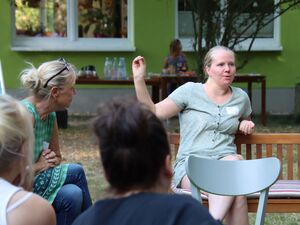 Junge Frau sitzt auf einer Holzbank. Sie hebt den rechten Arm. Frau frägt ein kurzes mintgrünes Kleid und hat die blonden Haare sträng nach hinten gekämmt. Im Hintergrund ist ein kleines Büfett mit Getränken aufgebaut.