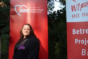 Portrait: Junge Frau mit langen braunen Haaren, zu zwei Zöpfen gebunden, sie trägt schwarze Kleidung. Sie steht vor einem roten AWO RollUp Banner.