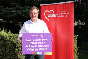 Portrait: Draußen. Mann in weißem Hemd und Jeans, er trägt eine schmale Brille. Er hält ein lila Schild und steht vor einem roten AWO RollUp Banner. Auf dem Schild steht: Rente muss für gutes Leben reichen - Strukturelle Kürzungen rückgängig machen.