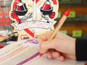 Runder Tisch auf dem eine Unterschriftenliste liegt. Person trägt sich in Liste ein. AWO Kugelschreiber, Becher und Tischaufsteller in Herzform auf dem Tisch.