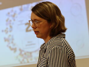 Frau steht im Veranstaltungsraum. Im Hintergrund die Präsentation an der Wand. Frau trägt Brille und eine gestreifte Bluse.