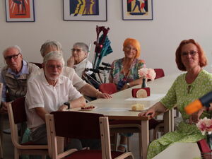 Raum mit Menschen an Tischen. Im Vordergrund rechts: Blonde Frau mit Mikro in der linken Hand.