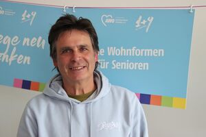 Portrait. Mann mit dunklen Haaren, grünen T-Shirt unter einem hellblauen Hoody. Er lächelt in die Kamera.