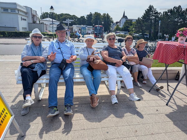 Sonnig, 4 Menschen sitzen auf einer weißen Bank, einige tragen Sonnenhüte. Zwei weitere Menschen sitzen auf einer niedrigen Mauer. Dahinter Straßen und Gebäude.