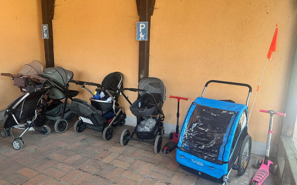 Kinderwagen Parkplatz mit Schild dafür gekennzeichnet. 5 Kinderwägen, ein Kinderfahrradanhänger und 2 Kinderroller parken dort.