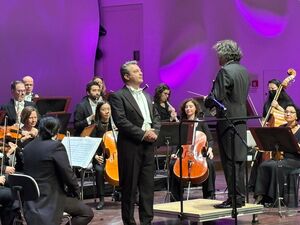 Orchester mit Dirigent und einem Tenor auf der Bühne. Alle schwarz gekleidet. Männer in Anzug und weißem Hemd und weißer Fliege.