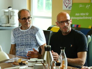 Zwei Männer sitzen nebeneinander am Tisch. Rechts der Mann mit Brille und dunklem T-Shirt spricht. Hinter ihm ein grünes Schild mit Text: Mehr Mittel für Wohnungsbauförderung.