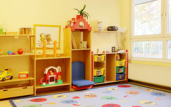 Heller Raum, Regal mit Spielzeug steht an einer gelben Wand, davor ein Teppich mit bunten Buchstaben-Luftballons.