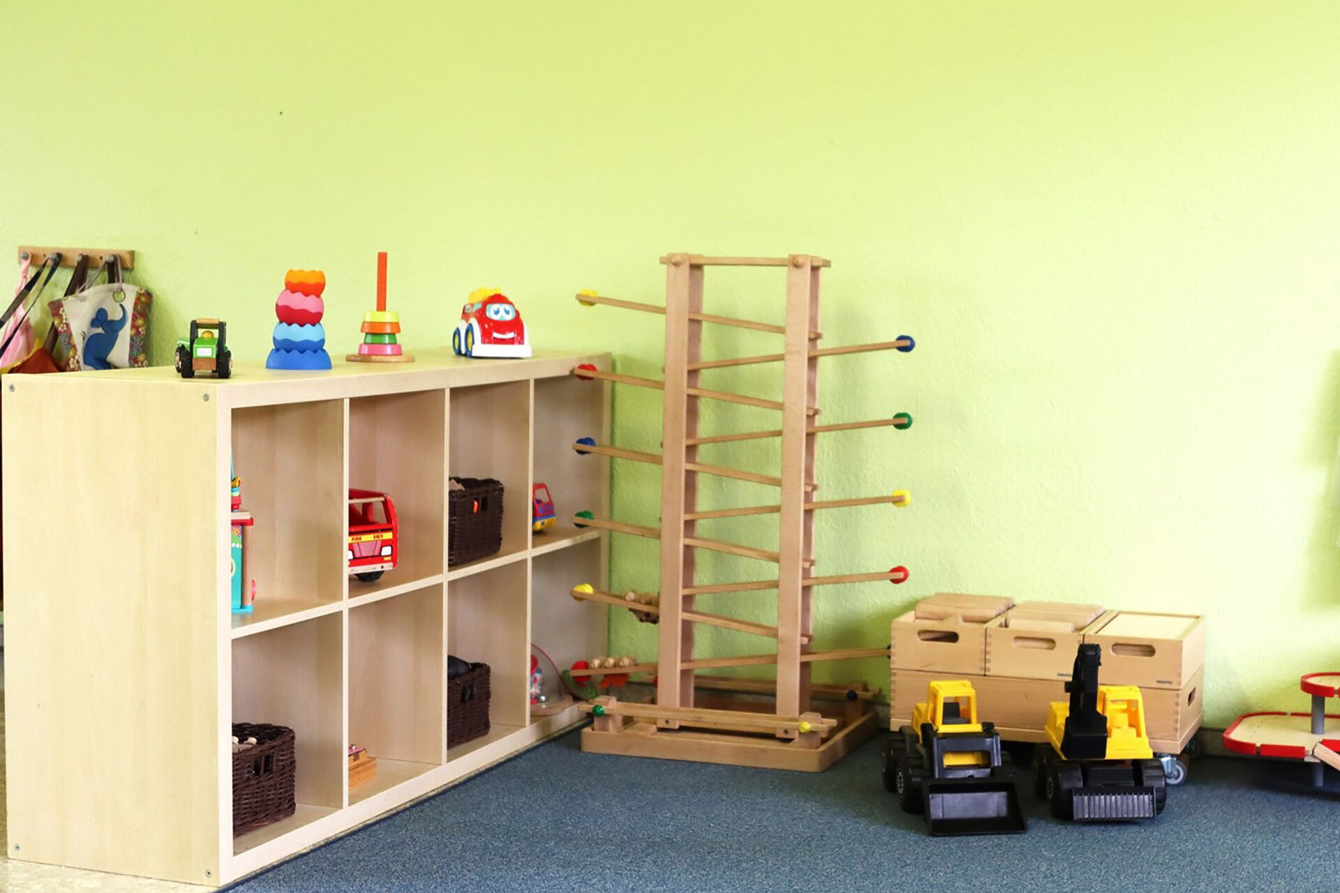 Spielzimmer mit Spielsachen. Ein Regal mit Feuerwehrauto, Trecker, Stapelturm. Auf dem Fußboden stehen eine Holzkugelbahn und zwei gelbe Bagger.