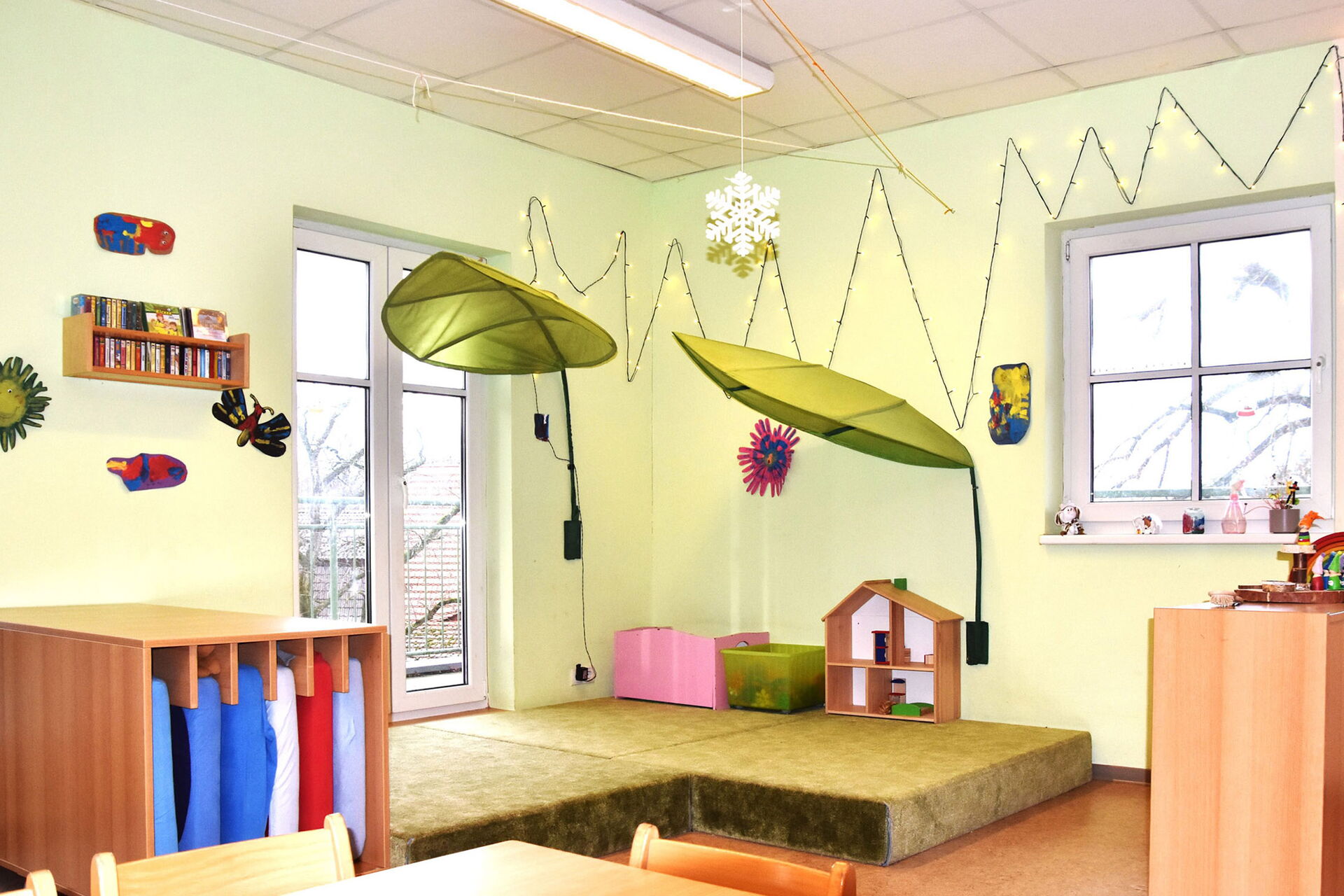 Spielzimmer. Ruhebereich. In einem Regal sind Matratzen. Ein Liegebereich mit 2 großen grünen Lampen darüber, die wie Blätter aussehen.